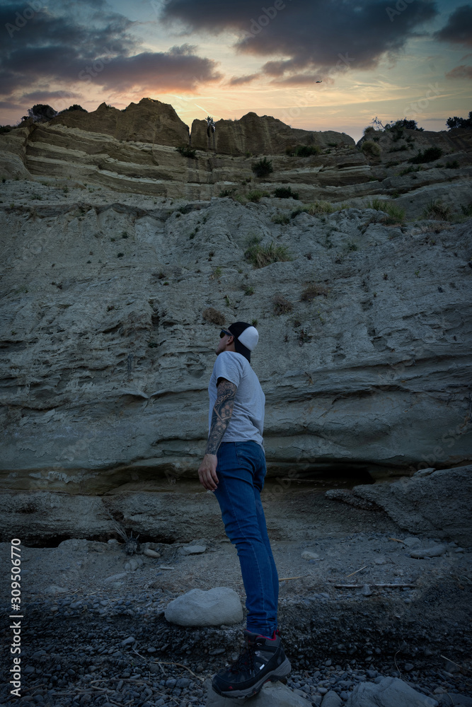 Young man wearing a cap posing in profile while looking at high cliffs, a big wall of rock and clay. Beautiful sunset scene. Vertical photography