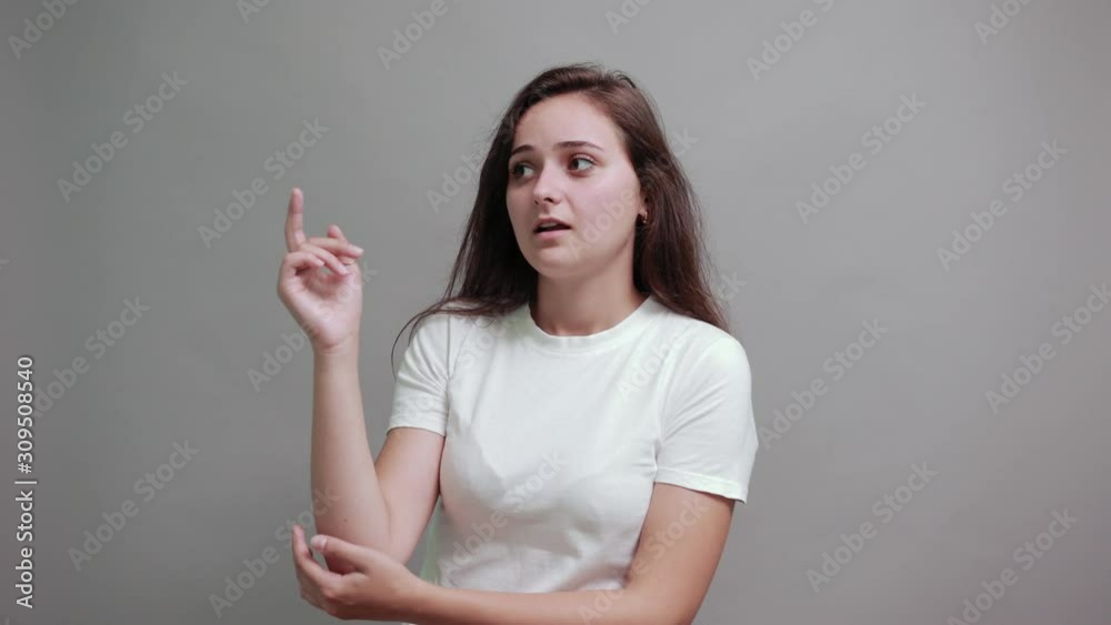 Charming caucasian young woman in fashion white shirt pointing finger aside, keeping hand on elbow isolated on gray background in studio. People sincere emotions, lifestyle concept. Charming caucasian young woman in fashion white shirt pointing finger aside, keeping hand on elbow isolated on gray background in studio. People sincere emotions, lifestyle concept.