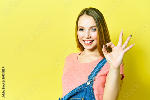 Funny and joyful girl shows okay sign and winks looking at camera isolated on yellow background.