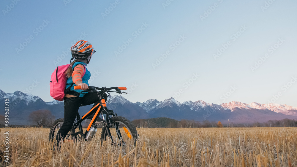 One caucasian children walk with bike in wheat field. Little girl walking black orange cycle on background of beautiful snowy mountains. Biker stand with backpack and helmet. Mountain bike hardtail. One caucasian children walk with bike in wheat field. Little girl walking black orange cycle on background of beautiful snowy mountains. Biker stand with backpack and helmet. Mountain bike hardtail.