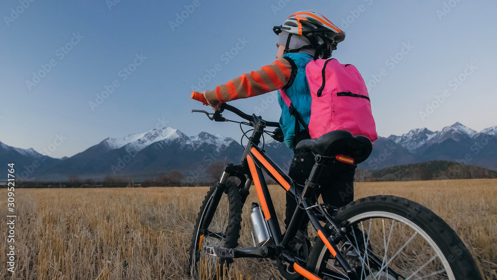 One caucasian children walk with bike in wheat field. Little girl walking black orange cycle on background of beautiful snowy mountains. Biker stand with backpack and helmet. Mountain bike hardtail.