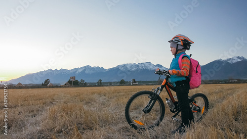 One caucasian children walk with bike in wheat field. Little girl walking black orange cycle on background of beautiful snowy mountains. Biker stand with backpack and helmet. Mountain bike hardtail.