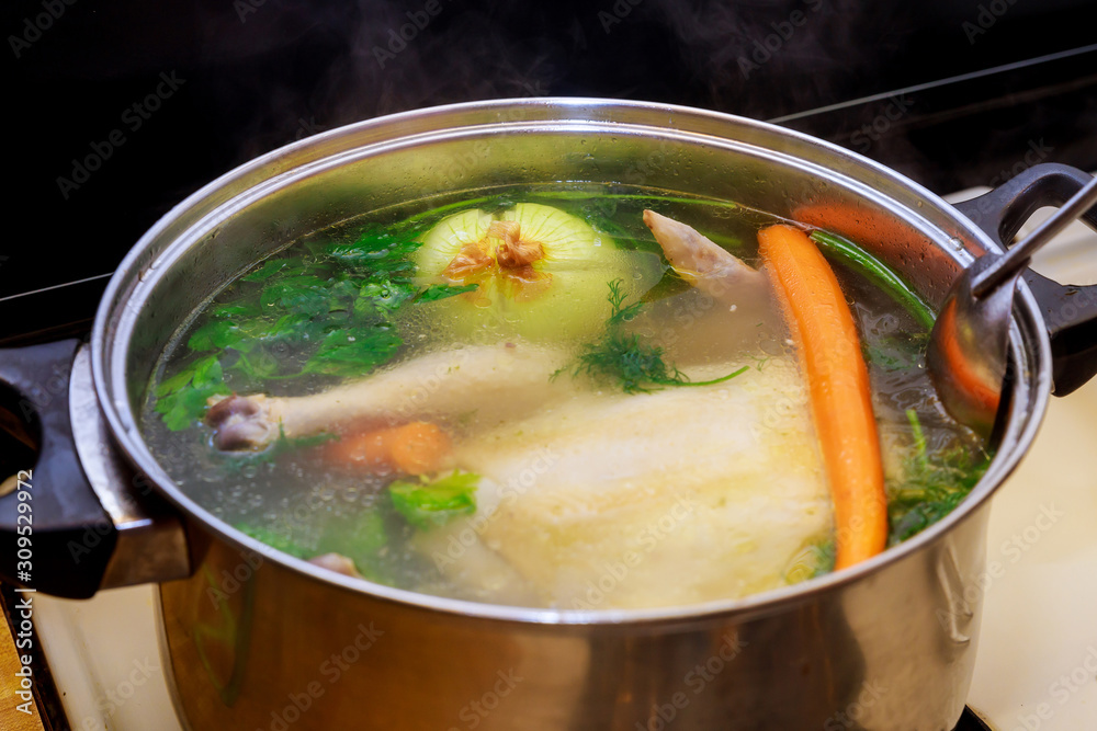 Chicken broth with carrots, whole chicken, onion, celery and parsley in steel pot.