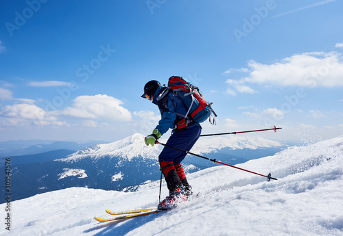 Sportsman skier in bright clothing, helmet and goggles with backpack riding down steep snowy slope on copy space background of blue sky and mountain landscape. Winter holidays, extreme sport concept.