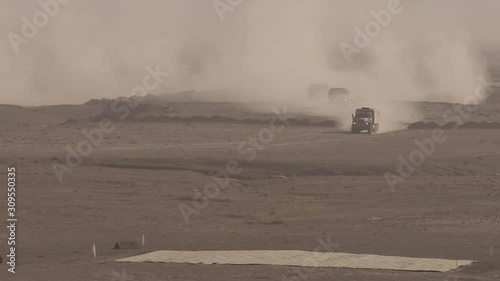 Military cargo convoy rides fast in desert sand dunes, leaving behind a massive trail of dust, military operation. The convoy group is moving in formation to meet the combat attack