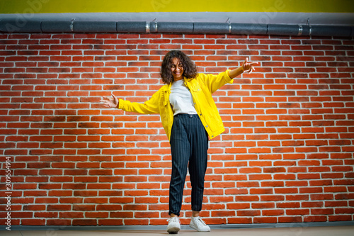 Lovely slim girl in yellow jacket posing with sincere smile. Pretty curly haired female model dancing with hands streight on brick wall background. photo