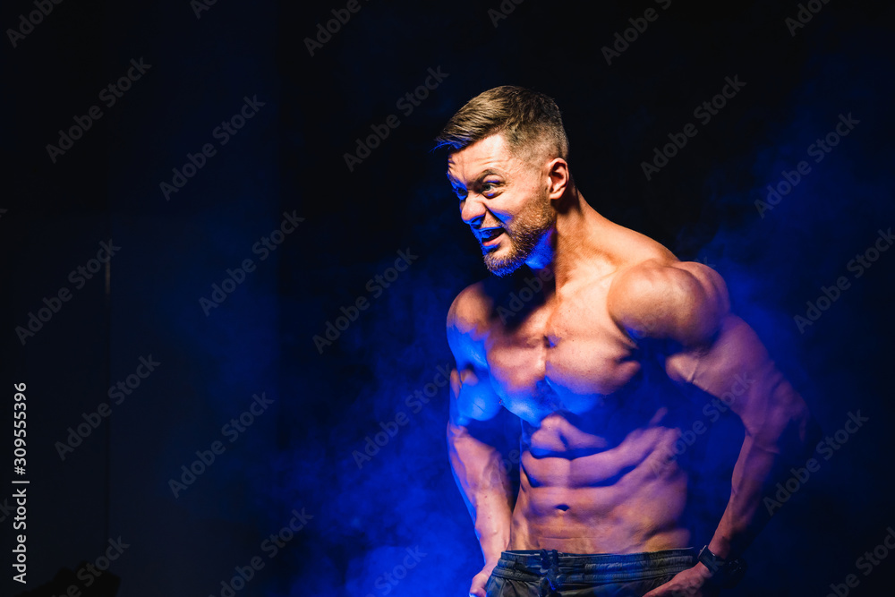 Strong brutal bodybuilder man with perfect abs, shoulders,biceps, triceps and chest showing his biceps. Scream motivation. Blue filter on dark background