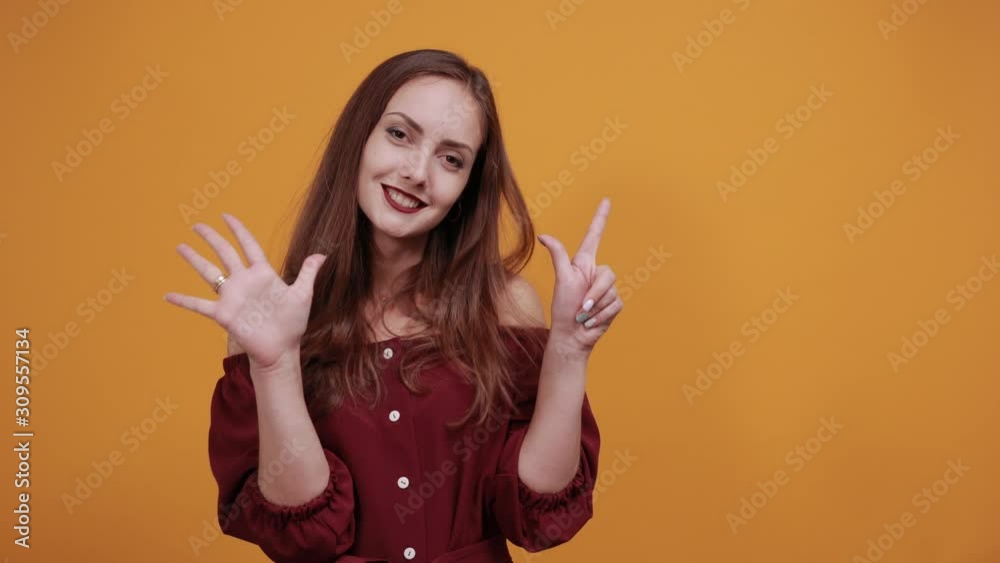 Cheerful caucasian woman in maroon dress isolated on orange background in studio showing seven fingers, smiling. People sincere emotions, lifestyle concept. Cheerful caucasian woman in maroon dress isolated on orange background in studio showing seven fingers, smiling. People sincere emotions, lifestyle concept.