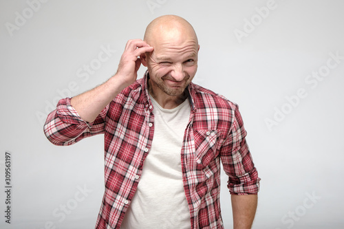 Tableau sur toile Bald man with stubble on face, looks into the camera with repentance, smiles guiltily and narrows one eye