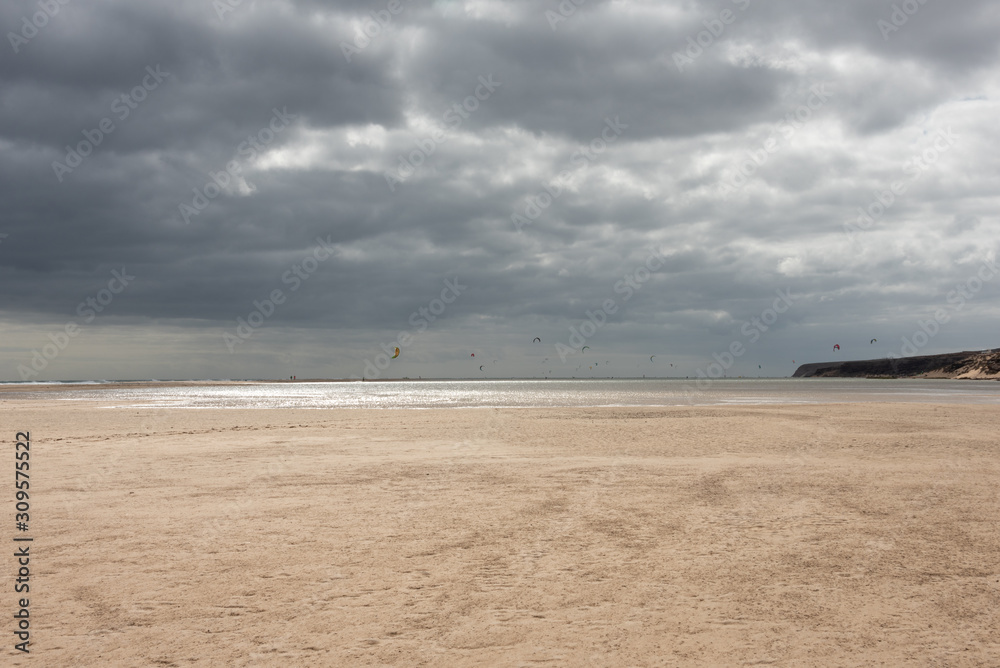 Playa de Sotavento - wide beach during low tide under cloudy sky. Kite surfers in the background. Costa Calma, Fuerteventura, Canary Islands, Spain. Playa de Sotavento - wide beach during low tide under cloudy sky. Kite surfers in the background. Costa Calma, Fuerteventura, Canary Islands, Spain.
