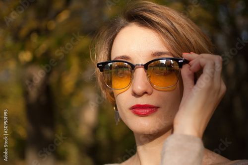 Young and beautiful girl with short hair and yellow sunglasses with mirror surface is posing against the trees with leaves background while spending time in the autumn park