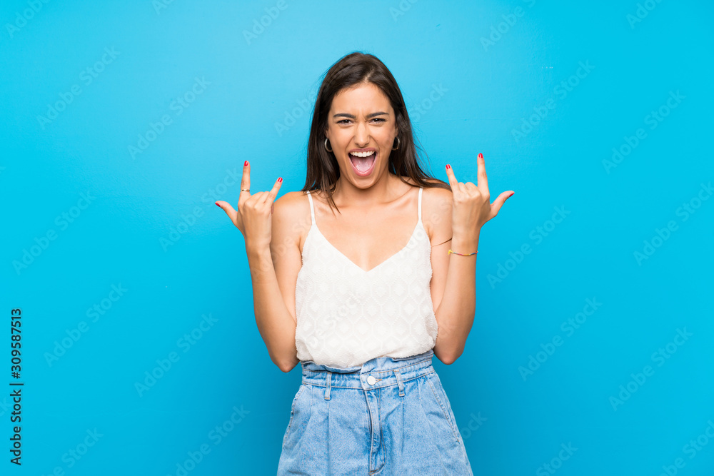 Young woman over isolated blue background making rock gesture Young woman over isolated blue background making rock gesture