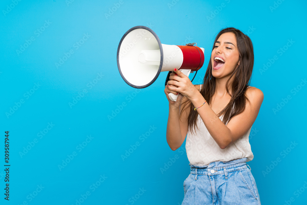 Young woman over isolated blue background shouting through a megaphone - obrazy, fototapety, plakaty 