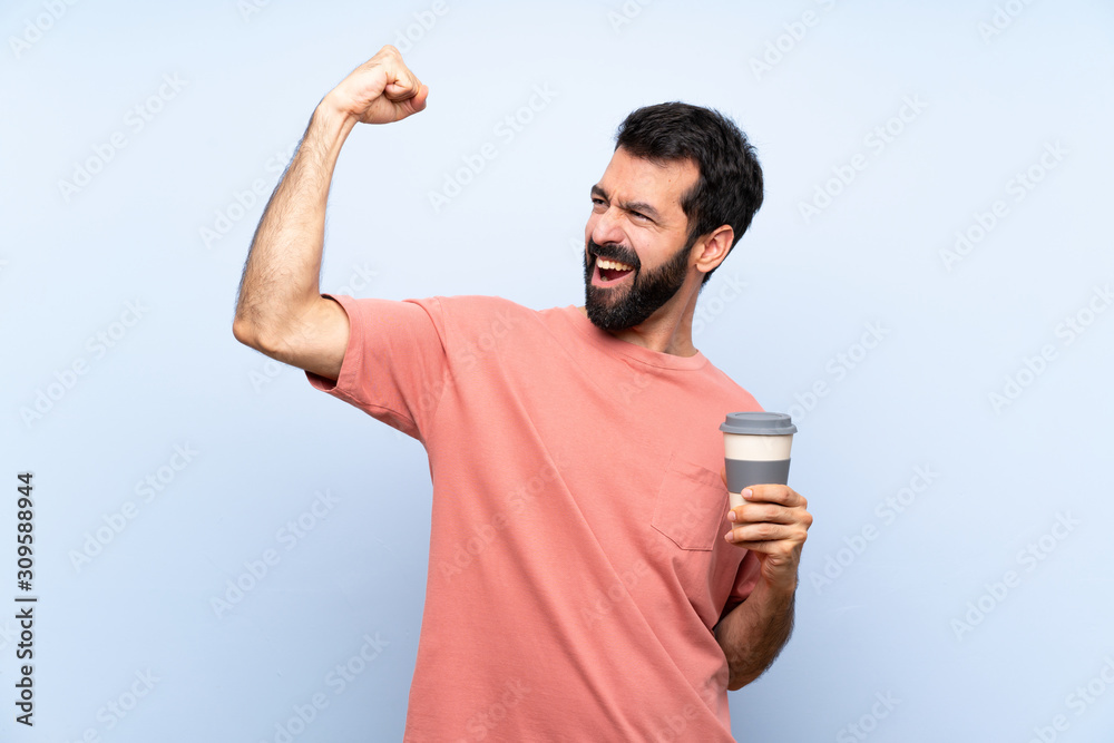 Young man with beard holding a take away coffee over isolated blue background celebrating a victory Young man with beard holding a take away coffee over isolated blue background celebrating a victory
