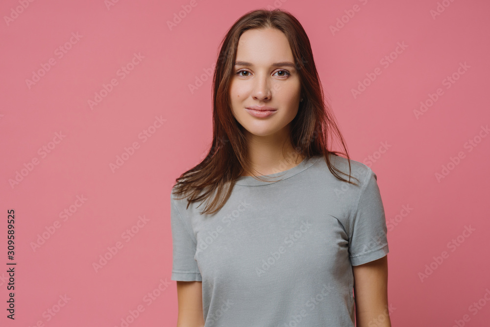 Obraz premium Adorable millennial girl with straight dark hair, wears casual t shirt, looks seriously at camera, has healthy skin, poses over pink background. Pleasant looking woman has talk with someone.