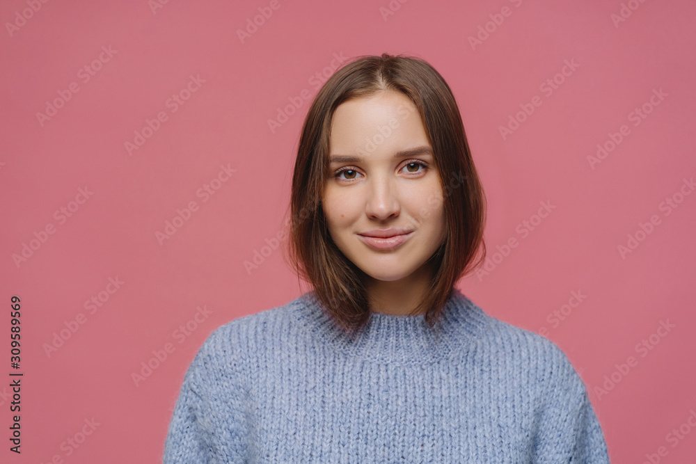 Beautiful adult woman with dark hair, has calm face expression, wears knitted sweater, poses at studio against pink background, thinks about something pleasant during winter time, has tender look Beautiful adult woman with dark hair, has calm face expression, wears knitted sweater, poses at studio against pink background, thinks about something pleasant during winter time, has tender look