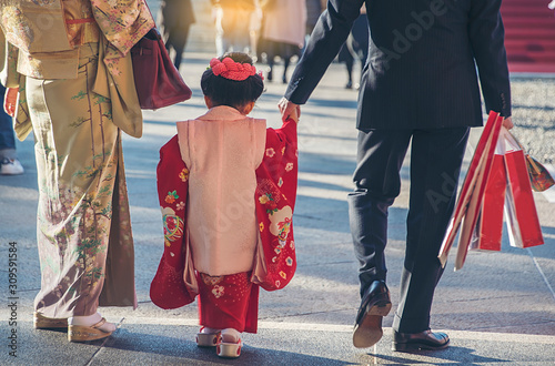 The mother and daughter wear kimono as a national dress in Japan, walk back from the temple and father wear a suit, carry a shopping paper bag. S