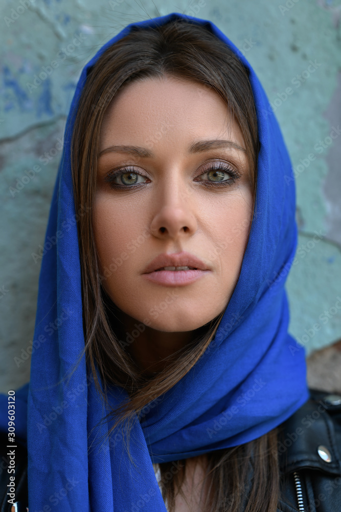 Close up portrait of young religious brunette hair green gentle eyes beautiful face adult woman or young girl with blue silk scarf covered head in black leather jacket on old brick wall background Close up portrait of young religious brunette hair green gentle eyes beautiful face adult woman or young girl with blue silk scarf covered head in black leather jacket on old brick wall background