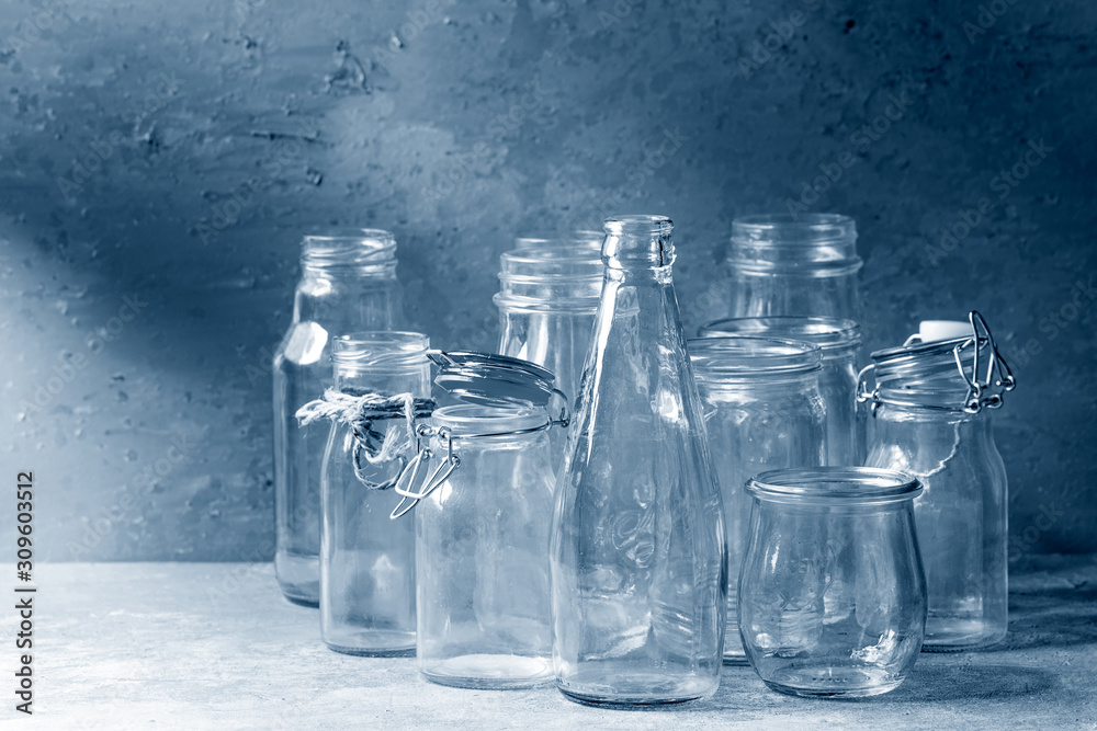Fototapeta Variety of different shape empty opened glass bottles with and without lids standing on grey table with wall as background. Color of the year 2020 classic blue toned