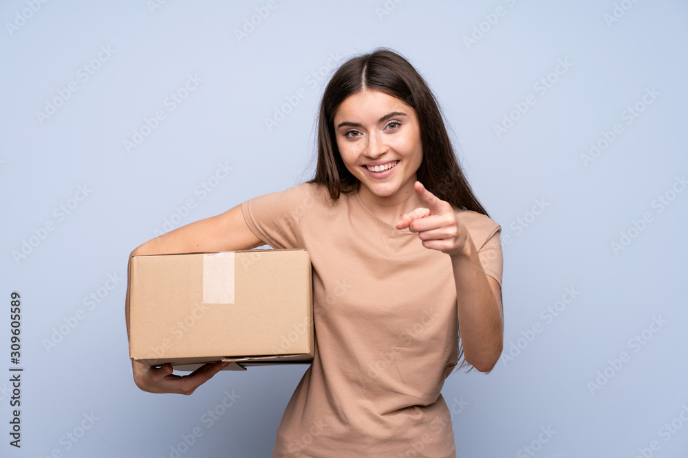 Young woman over isolated blue background holding a box to move it to another site while pointing to the front Young woman over isolated blue background holding a box to move it to another site while pointing to the front