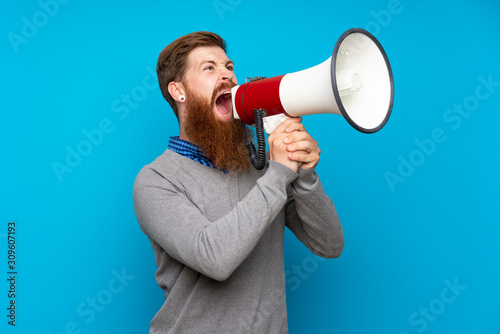 Redhead man with long beard over isolated blue background shouting through a megaphone