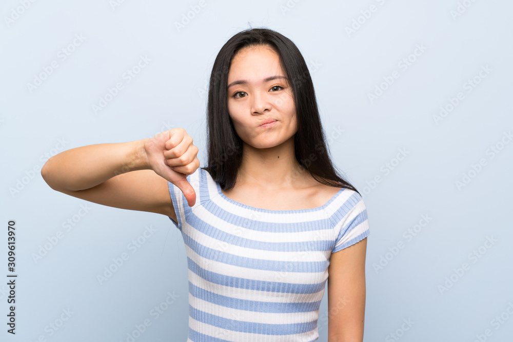 Teenager asian girl over isolated blue background showing thumb down sign Teenager asian girl over isolated blue background showing thumb down sign