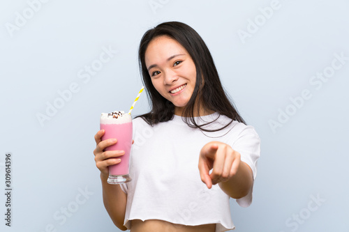 Teenager asian girl holding a strawberry milkshake points finger at you with a confident expression