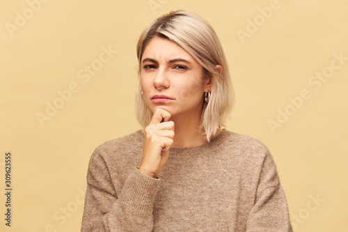 Beautiful fashionable young suspicious European female in cashmere pullover holding hand on her chin and staring at camera with suspicion and mistrust, screwing up eyes. Human facial expressions