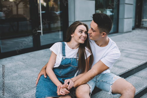 Positive male and female enjoying date time at urban setting embracing each other and looking in eyes, happy hipster guys communicating and smiling, concept of warm feelings and relationship