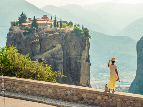 Young woman with white dress and large hat standing on wall in front of greece meteor mountains, monastery and village in the background photo