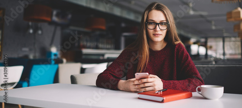 Portrait of young attractive hipster girl in eyeglasses looking at camera holding cellular, beautiful female student sitting at cozy cafeteria using mobile phone and chatting with friends online © GalakticDreamer