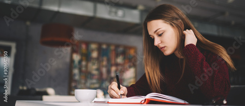 Attractive young talented writer 20 years old creating new book atricles while sitting at cafeteria, thoughtful student preparing for important college exam writing answers for question in notebook photo