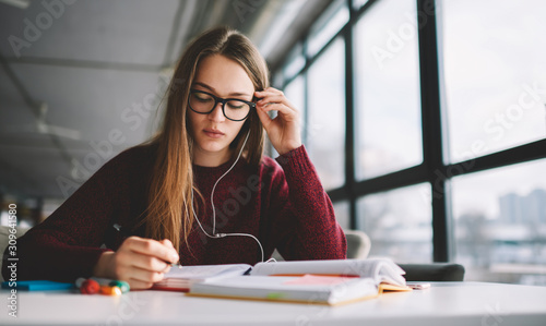 Pensive female in eyeglasses creating new plan of organisation work at cafeteria while enjoying new music playlist and listening radio, concentrated student doing homework indoors in earphones