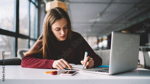 Young attractive female writer scribing notes to textbook prepare for exam, girl doing homework sitting desktop with modern laptop computer, woman making plan for tomorrow and creating to do list