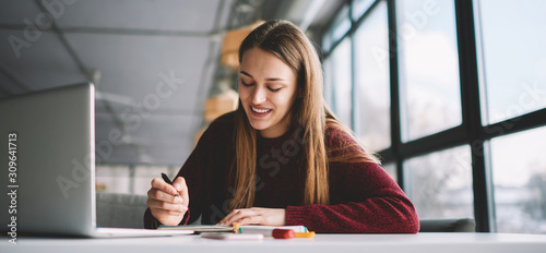 Smiling attractive hipster girl doing homework checking information using modern laptop computer, skilled happy female student writing creative ideas to notebook sitting in university campus