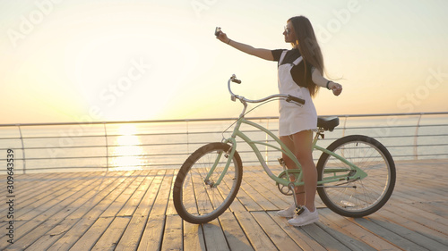 Young attractive woman uses a smartphone and riding vintage bike near the sea during sunrise or sunset