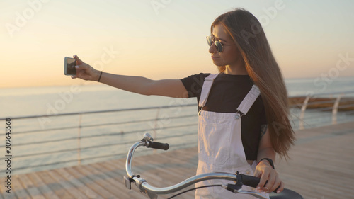 Young attractive woman uses a smartphone and riding vintage bike near the sea during sunrise or sunset