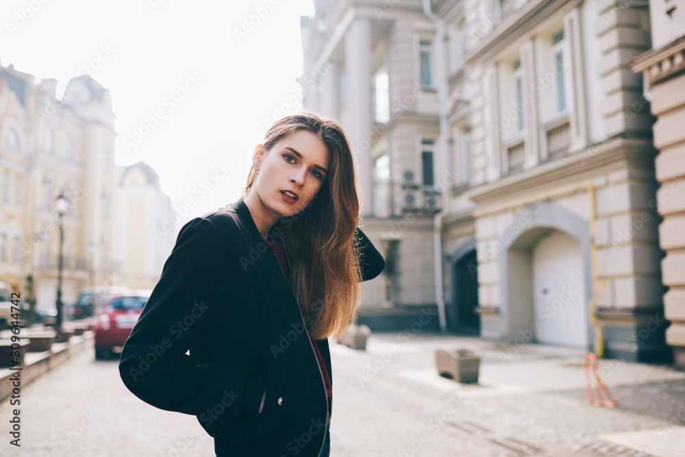 Portrait of attractive woman in stylish black jacket looking at camera strolling, young hipster girl spending time outdoors while walking at city on sightseeing tour during spring weekends