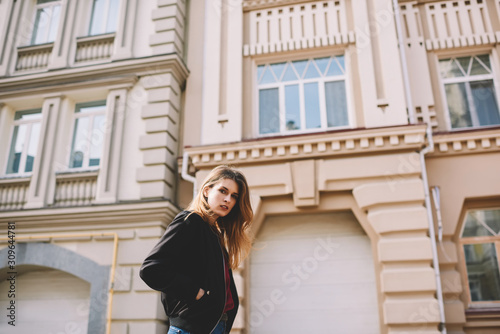 Portrait of beautiful female journalist in black jacket resting time while walking on street looking at camera, attractive stylish hipster girl 20 years old strolling outdoors on urban setting