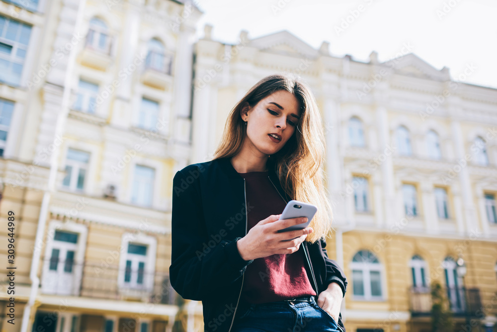 Fototapeta premium Hipster girl sending text messages and searching information about touristic route via cellphone outdoors, beautiful trendy woman chatting with friends online in social networks using modern telephone