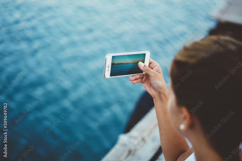 Back view of female holding mobile phone and taking nature pictures with camera on modern smartphone, cropped photo of teenage hipster girl using advanced mobile phone for making video for new blog