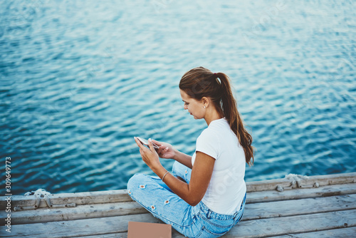 Attractive hipster girl outdoors check mail by new cellular phone with 4g connection on pier, back view of positive woman sending text messages on modern mobile phone while resting near river