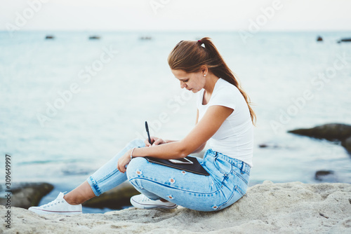 Charming teenage hipster girl in denim jeans writing interesting review about travel in notebook outdoors, young fashionable beautiful woman enjoying drawing sketch nature near calm lake surface