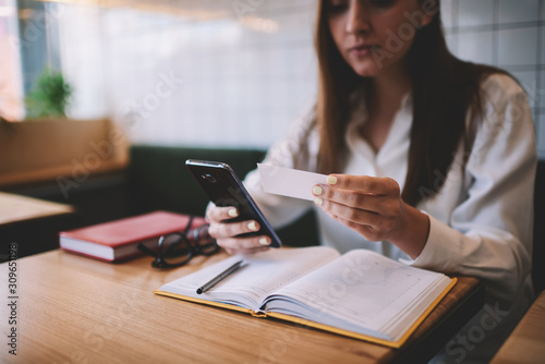 Young hipster girl 20s holding smartphone in hand and looking at business card for dialing colleague for discussing meeting, female student writing email via modern mobile phone while sitting indoors