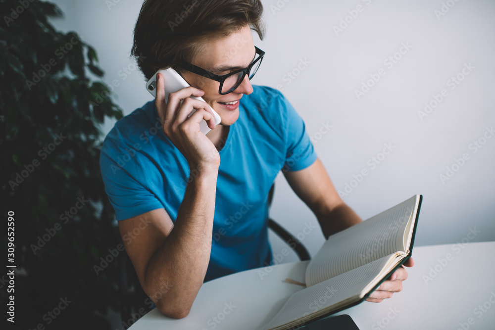 Happy cheerful hipster guy in optical glasses checking notes information from personal planner during smartphone conversation with best friend discussing time of meeting, millennial man phoning