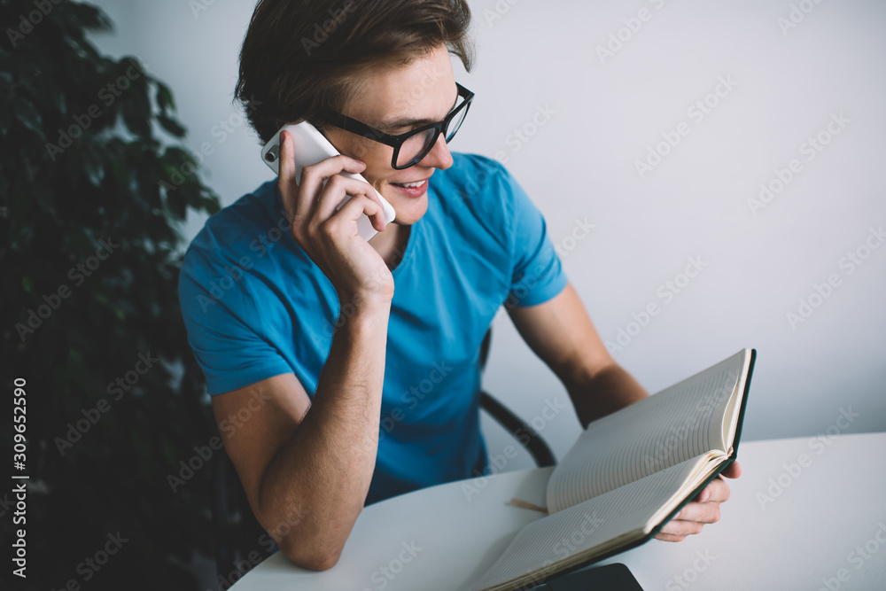 Happy cheerful hipster guy in optical glasses checking notes information from personal planner during smartphone conversation with best friend discussing time of meeting, millennial man phoning Happy cheerful hipster guy in optical glasses checking notes information from personal planner during smartphone conversation with best friend discussing time of meeting, millennial man phoning