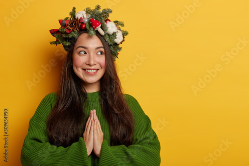Religious pretty Asian woman keeps palms together in prayig gesture, looks aside, wears Christmas wreath, green jumper, believes in good fortune, isolated over yellow wall copy space on left