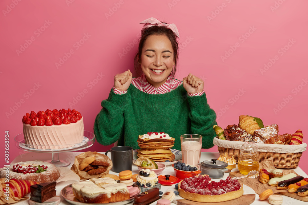 Joyous female cheers seeing many desserts, clenches fists, poses at table with tempting seductive homemade sweets, smiles happily, wears winter jumper, isolated over pink wall. Tasting yummy treat Joyous female cheers seeing many desserts, clenches fists, poses at table with tempting seductive homemade sweets, smiles happily, wears winter jumper, isolated over pink wall. Tasting yummy treat