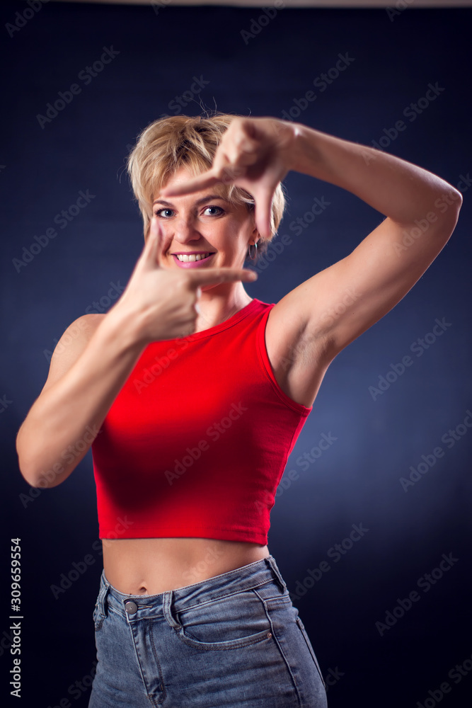 A portrait of young woman with short blond hair in red shirt showing photo frame gesture in front of black background. Lifestyle, emotions and beauty concept A portrait of young woman with short blond hair in red shirt showing photo frame gesture in front of black background. Lifestyle, emotions and beauty concept