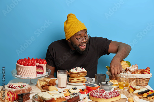 Cheerful unshaven man points at delicious fancy cake  likes eating pastry  poses at blue background near table with freshly baked confectionery  wears stylish yellow hat  black t shirt. Sweet delicacy